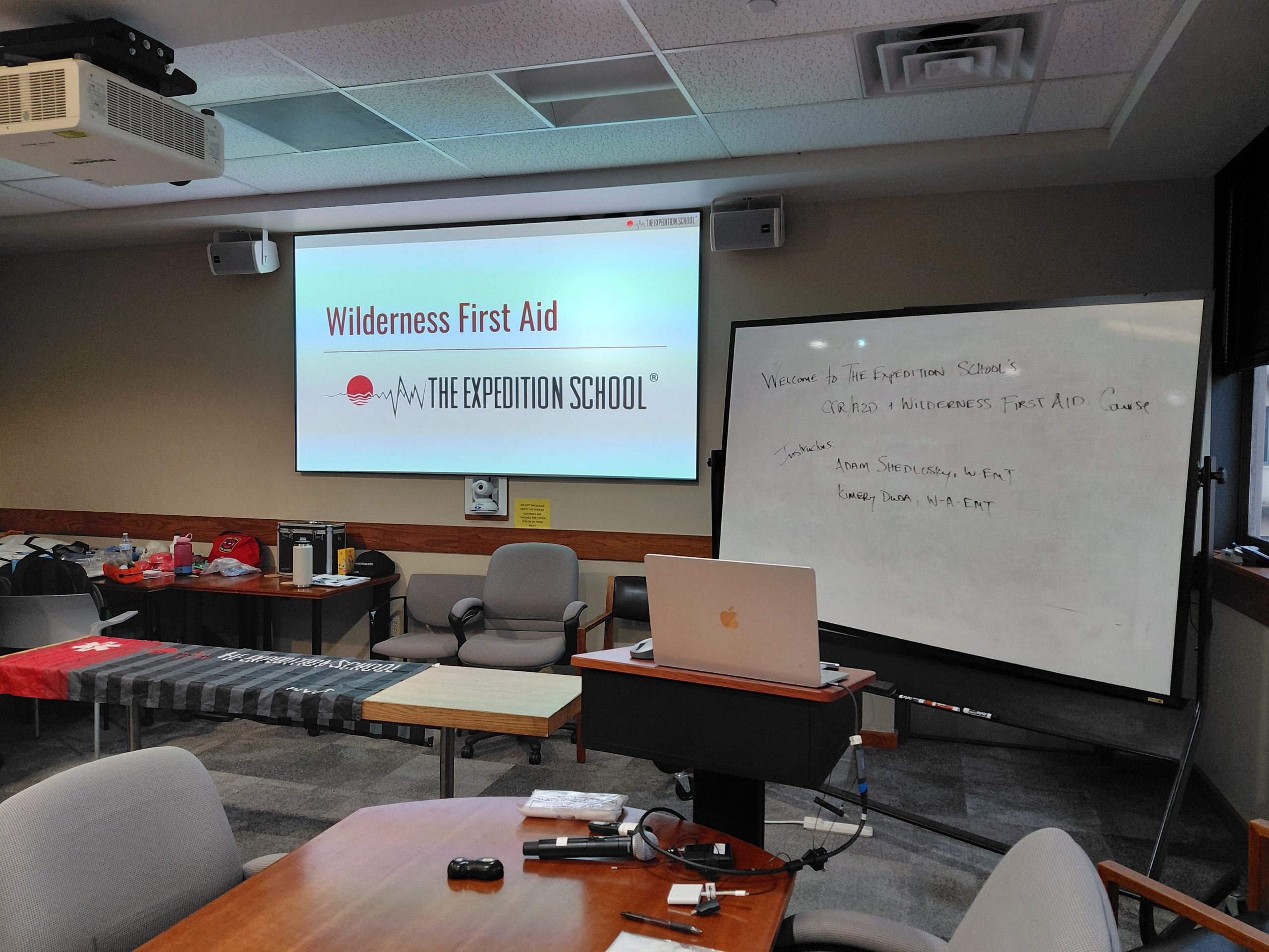 Empty classroom at UT Austin before the Expedition School Wilderness First Aid course begins, showing the opening slide on screen and a whiteboard listing instructors Kimery Duda and Adam Shedlosky.