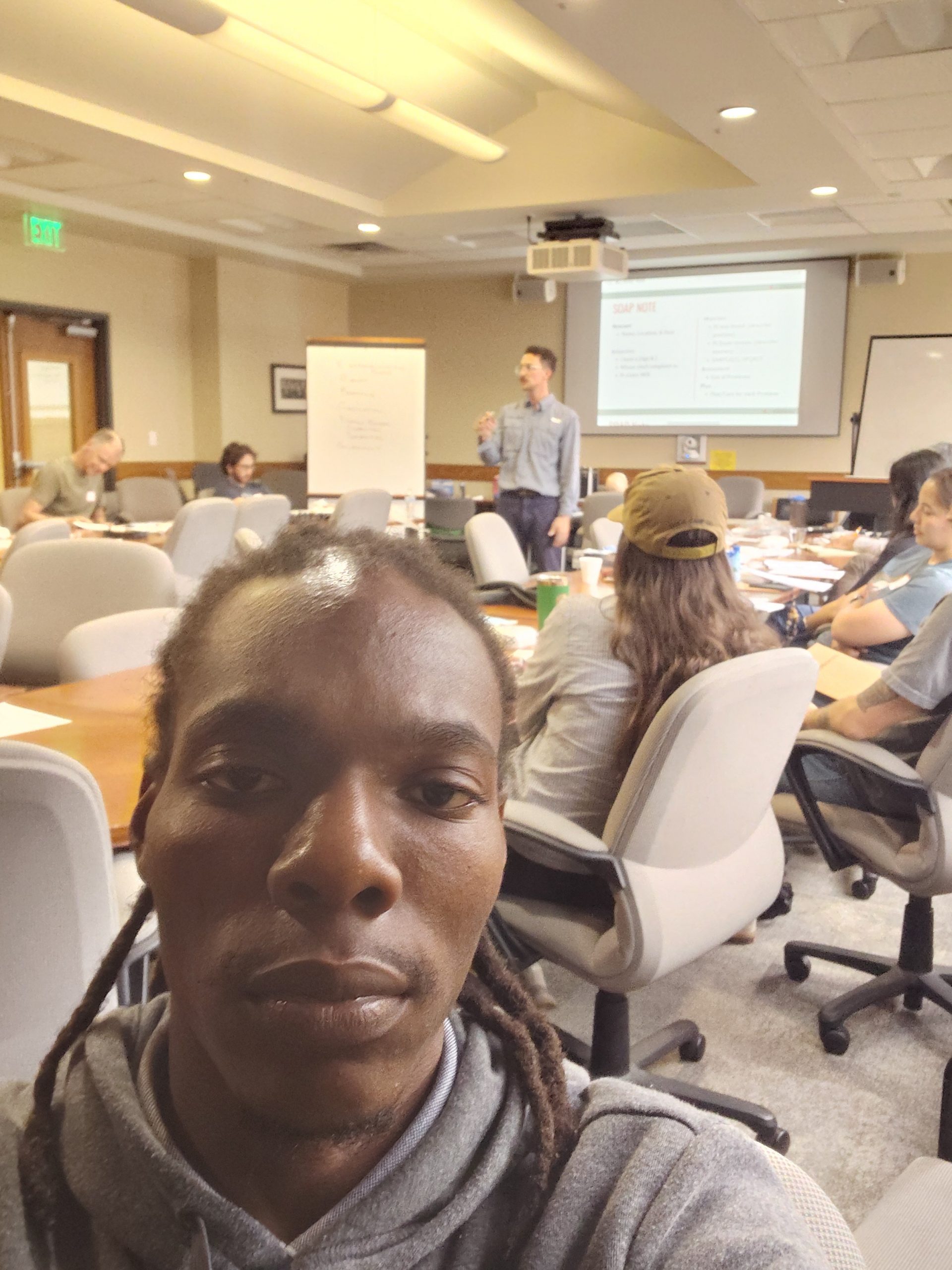 Mitchel Monsels, YLAI fellow from Suriname, takes a selfie during the Expedition School Wilderness First Aid course at UT Austin, with instructor Adam Shedlosky presenting the SOAP note framework visible in the background.